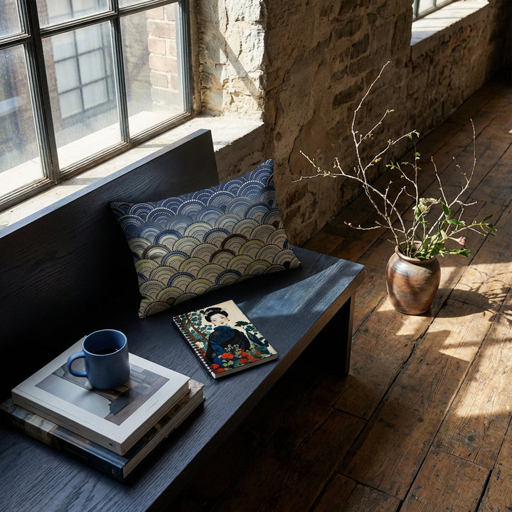 Photographie d’intérieur ambiance zen loft moderne, gros plan sur une banquette en bois  orné d'un coussin et d'un carnet mzelle design, fenêtre industrielle, mur brut en vieilles pierres, sol en bois , tasse de café en céramique  sur une pile de livres d'art, branche ikébana dans un pot au sol. 