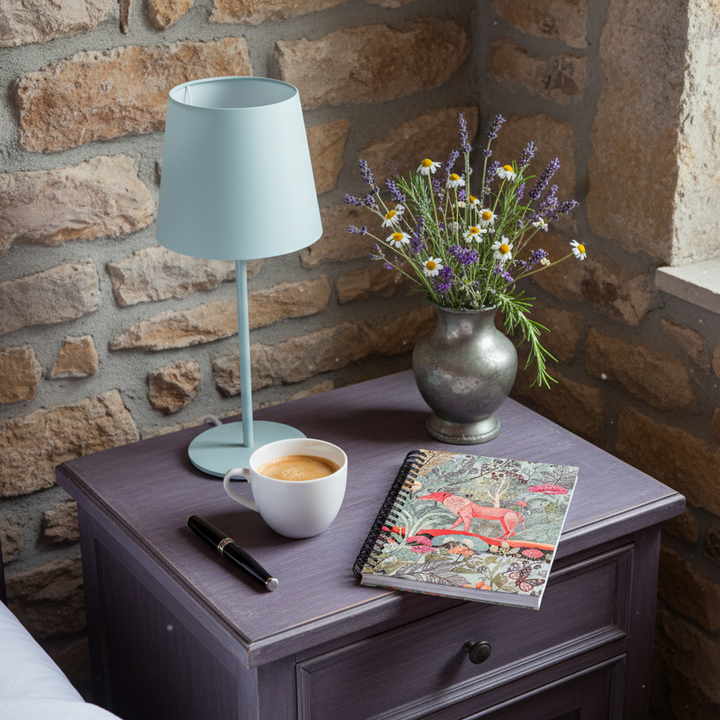 Photo professionnelle décoration de d'une chambre d'hôte en maison de campagne provençale, vue sur une table de chevet en bois peint violet foncé avec un carnet mzelle design posé en évidence, une tasse de café, une lampe moderne bleu glace, un stylo plume, Un vase en étain de fleurs sauvage, mur en pierres brutes et fenêtre.
