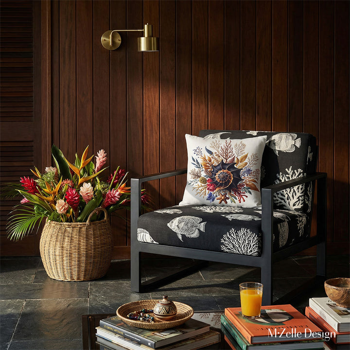 Photographie intérieure de style éditorial d’une villa créole d'architecte, vue de face d'un fauteuil épuré structure épaisse en métal noir avec des motif blanc représentant des poissons tropicaux et des coraux, orné d'un coussin carré motif coquillage, placé contre un mur de panneaux de bois sombre.