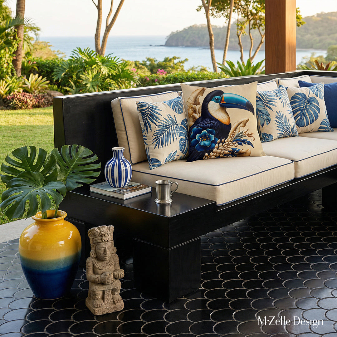 Photographie lifestyle extrêmement détaillée esprit estival, vue de face en gros plan sur un canapé en bois peint noir laqué, structure épaisse épurée qui comporte une extension pour faire une table basse intégrée, assises écrues à motifs graphiques de feuilles tropicales bleus, orné de grand coussin motif toucan, jardin et mer en arrière plan.