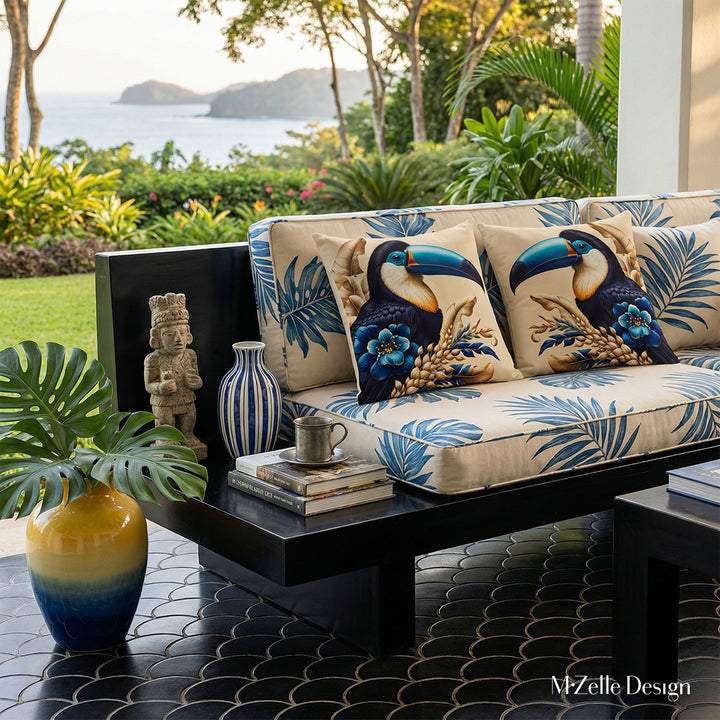 Photographie lifestyle extrêmement détaillée esprit estival, vue de face en gros plan sur un canapé en bois peint noir laqué, structure épaisse épurée qui comporte une extension pour faire une table basse intégrée, assises écrues à motifs graphiques de feuilles tropicales bleus, orné de grand coussin motif toucan, jardin et mer en arrière plan.