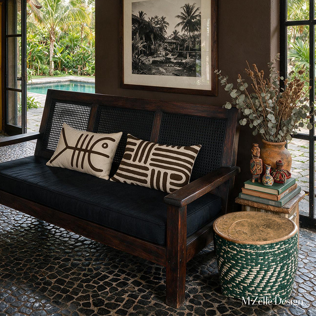 Photographie d’intérieur de style éditorial décoration d'hacienda modernisée. Canapé créole foncé au dossier en wengé et cannage noir, orné de deux coussins mzelle design motif tribal. Adossé à un mur peint en marron foncé uni sur lequel est accrochée une grande photographie en noir et blanc représentant un paysage exotique.