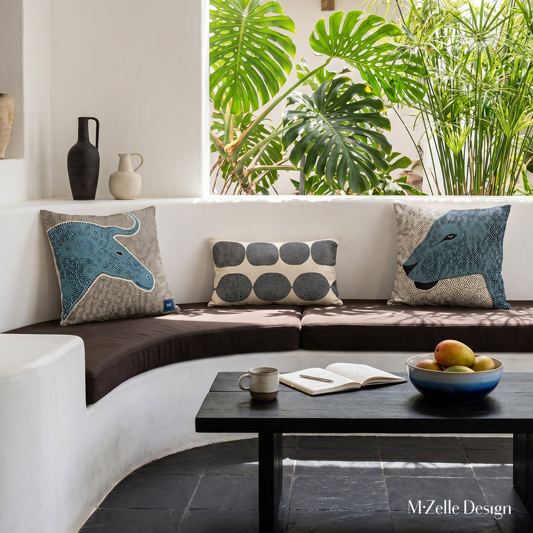 Photo professionnelle hautement détaillée, salon Arty dans une villa créole baignée de soleil avec murs blanchis à la chaux, canapé en demi-lune, intégré dans le mur , assises marron foncé avec trois coussins mzelle design style graphique, carrelage brutaliste noir au sol, Table basse en bois noir avec une tasse de café, carnet, coupe en grès bleu dégradé contenant des mangues. En arrière plan, une arche ouverte sur un jardin. 