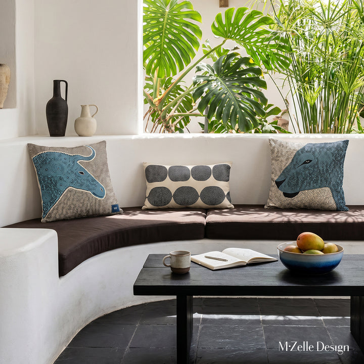 Photo professionnelle hautement détaillée, salon Arty dans une villa créole baignée de soleil avec murs blanchis à la chaux, canapé en demi-lune, intégré dans le mur , assises marron foncé avec trois coussins mzelle design style graphique, carrelage brutaliste noir au sol, Table basse en bois noir avec une tasse de café, carnet, coupe en grès bleu dégradé contenant des mangues. En arrière plan, une arche ouverte sur un jardin. 
