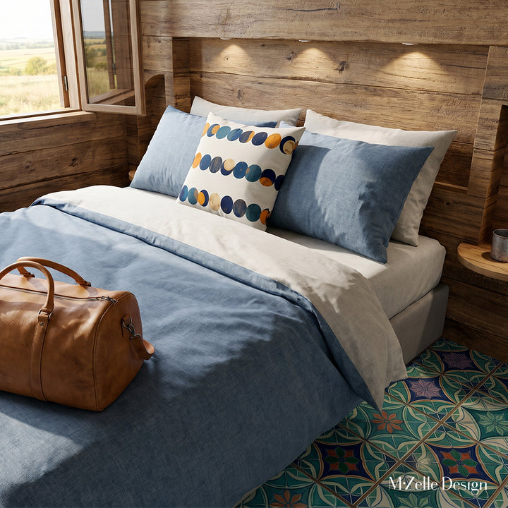 Photographie d’intérieur, chambre d'hôte rustique eco lodge minimaliste , vue en plongée en gros plan sur un coussin mzelle design motifs galets stylisés, posé sur un grand lit contre 2 oreillers, draps raffinés en coton de chambray bleu et écru, un sac de voyage en cuir camel sur le lit, mur en bois d'acacia brut et fenêtre ouverte, vue campagne, sol en carrelage élaboré spots lumineux, ambiance campagne.