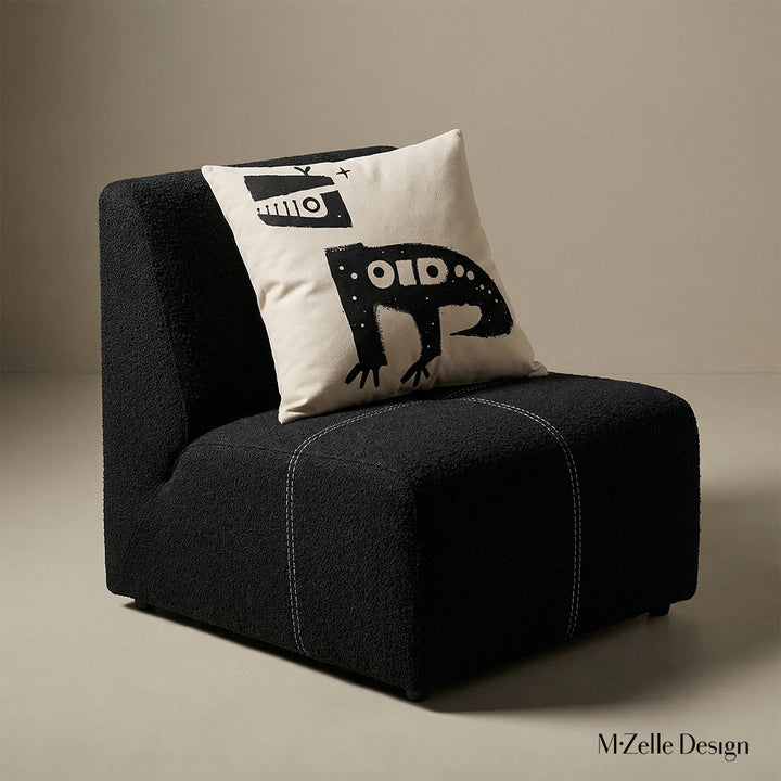 Photographie professionnelle  d'un fauteuil en en tissu laineux texturé noir, sans accoudoirs, assise surpiqué de fil blanc, orné d'un large coussin graphique mzelle design, disposé dans décor écru neutre. 