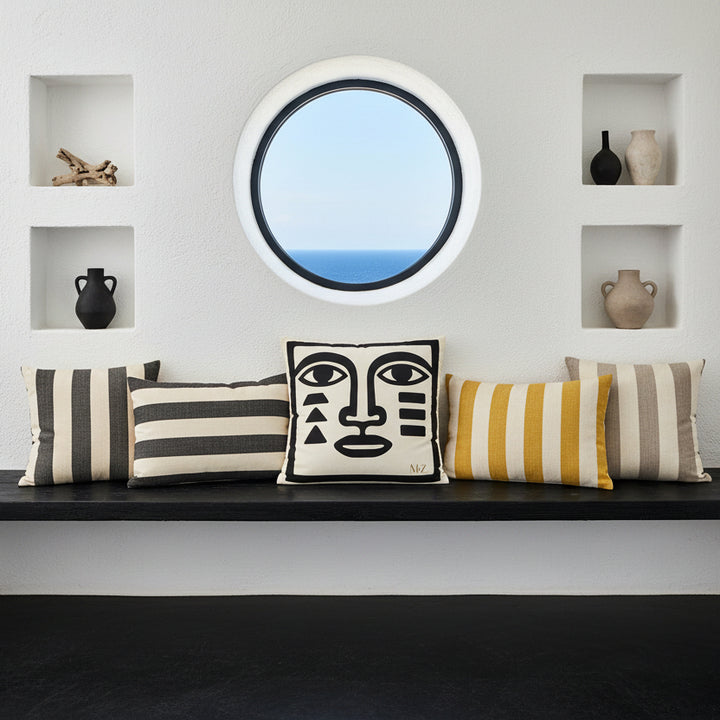 Photographie vue de face une banquette rustique en bois noir brut encastrée dans un mur de chaux blanc, orné au milieu le large coussin carré, et 3 coussins à rayures dans les tons noir, jaune et ecru, autour. atmosphère accueillante