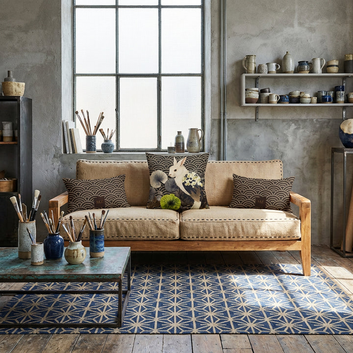 Photographie d’intérieur décoration de loft d’artiste céramiste, verrière industrielle, vue de face sur un canapé en bois de manguier, assises en toile de jute orné d'un large coussin mzelle design au centre, entouré de 2 coussins rectangles , tapis motif Kikkō noir et beige, objets artistiques (pinceaux dans un vase, livres d'art, rouleaux d'affiches dans un panier), étagère ouverte remplie de céramiques artisanales. 