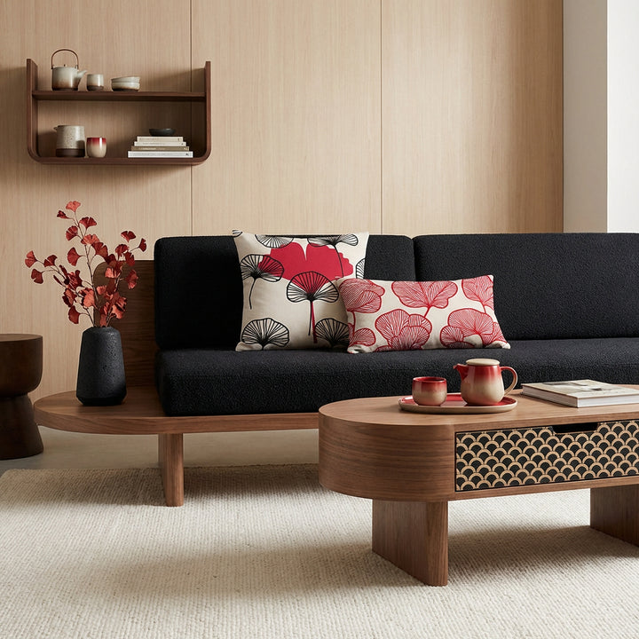 Photographie d'intérieur décoration japandi-nordique, épuré mais chaleureux, douce lumière naturelle. Vue de face d'un fauteuil-futon moderne assise en lin noir texturé orné d'un large coussin rectangle et carré, mzelle design motif ginkgo rouge et écru, extension en bois sur un côté du fauteuil servant de table basse, avec un tiroir. Service à thé en céramique à dégradé rouge et beige, livre.