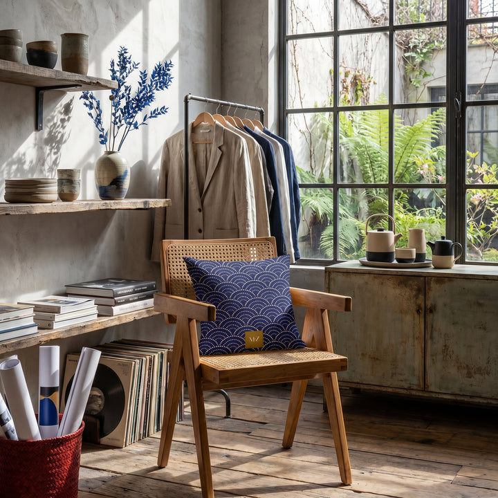 Photographie d’intérieur décoration de  boutique concept store, verrière industrielle, gros plan sur une chaise vue de face, en bois de manguier orné d'un coussin mzelle design motif japonais seigaiha, étagères de céramiques, vase avec des branches séchées ikebana, rangées de livres d'art, rouleaux d'affiches dans un panier, vêtements sur un portant.