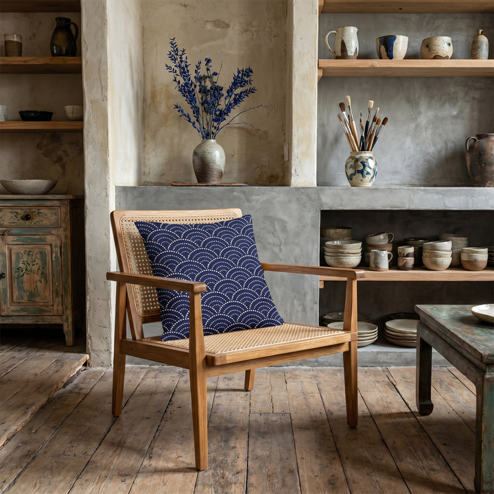 Photographie d’intérieur décoration de  boutique concept store, verrière industrielle, gros plan sur une chaise vue de face, en bois de manguier orné d'un coussin mzelle design motif bleu japonais seigaiha, étagères de céramiques, vase avec des branches séchées ikebana, rangées de livres d'art, rouleaux d'affiches dans un panier, vêtements sur un portant.