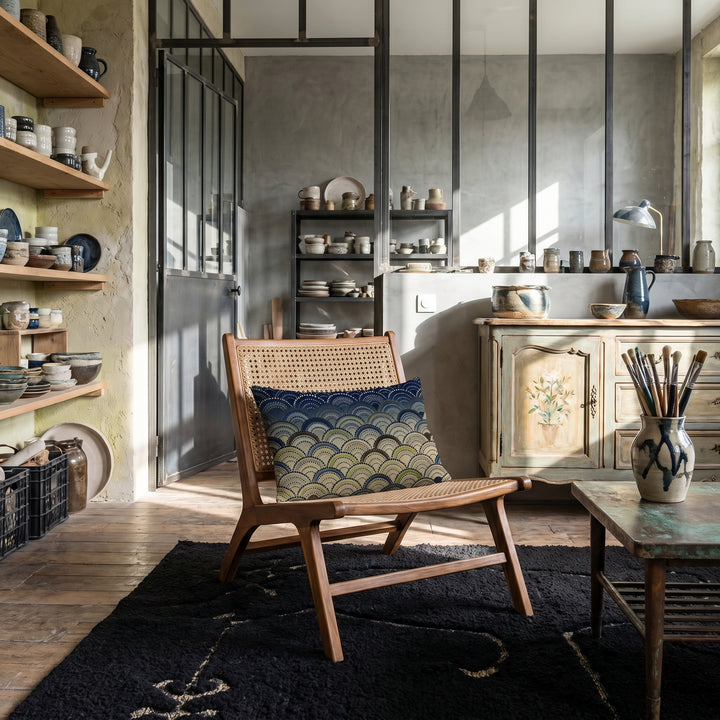 Photographie d’intérieur décoration de loft d’artiste céramiste,  gros plan sur une chaise vue de face, en bois de manguier et cannelage, orné d'un coussin mzelle design motif japonais seigaiha, tapis noir motif en laine bouclée, murs bruts enduit chaux et béton ciré gris, sol en bois ancien irrégulier. 