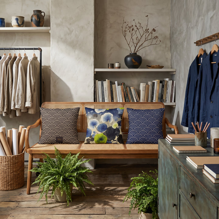 Photographie d’intérieur décoration de  boutique concept store, verrière industrielle, gros plan sur une banquette vue de face, en bois de manguier orné de trois coussins mzelle design motif japonais seigaiha et fleurs, étagères de céramiques, vase avec des branches séchées ikebana, rangées de livres d'art, rouleaux d'affiches dans un panier, vêtements sur un portant.