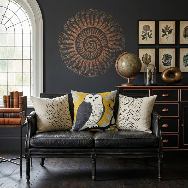 Photo professionnelle de décoration d'intérieur, salon esprit Wunderkammer.  Canapé en bois noir patiné vieilli, orné d'un large coussin mzelle design motif chouette,  entouré de chaque côté d'un coussin écru motif polka dots noir.  mur gris anthracite profond décoré d'un grand fossile stylisé cuivré au pochoir.