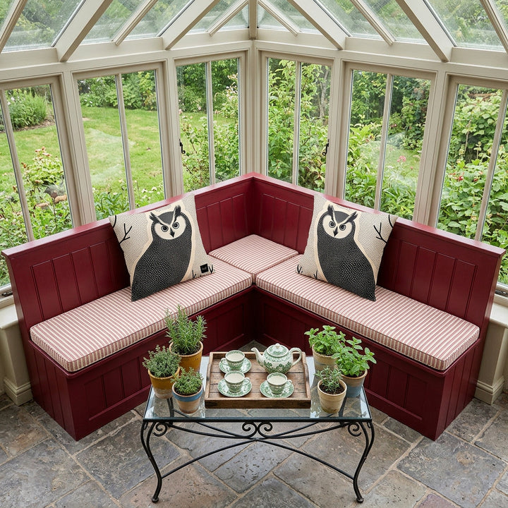 Photo professionnelle de décoration, verrière cottage anglais, vue en plongée d'une banquette d'angle en bois peint en rouge grenat, assises en twill à rayures fines rouge et écru , ornée de 2 coussins décoratifs mzelle design motifs hiboux de chaque côté. sol en carrelage rustique gris, table en fer forgé noir, garnie d'un service à thé en porcelaine vert et blanc dans un plateau en bois rustique,  plantes aromatiques. Atmosphère campagnarde.