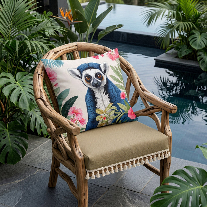 Photographie professionnelle d'un grand coussin mzelle design motif tropical lémurien, disposé sur une chaise au design contemporain en bois flotté,  assise beige finitions petits pompons écrus, placé devant une piscine sombre moderne à débordement et plantes exotiques. 