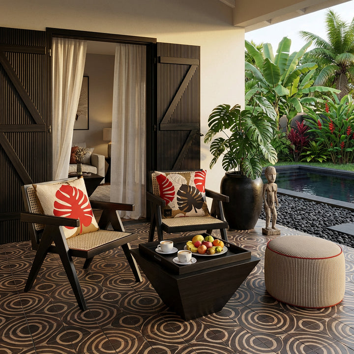 Photo de décoration de villa créole avec 2 sublimes fauteuils en ébène au design moderne, cannage beige orné chacun de coussin mzelle design, motif feuilles de monstera, table basse tasses de café et assiette de fruits, portes en bois peint rouge gravées  , carrelage géométrique de cercles, grand pot en grès noir contenant une plante monstera, jardin et piscine.