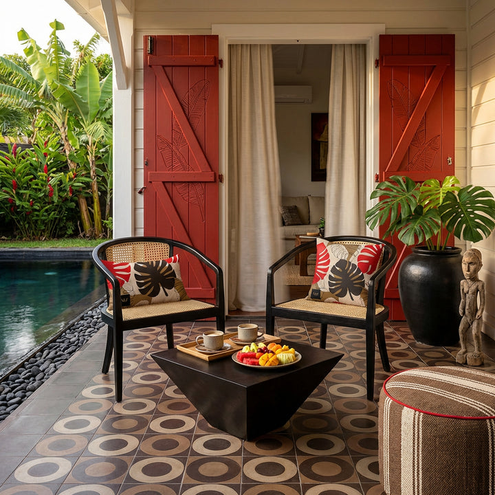 Photo de décoration de villa créole avec 2 sublimes fauteuils en ébène au design moderne, cannage beige orné chacun de coussin mzelle design, motif feuilles de monstera, table basse tasses de café et assiette de fruits, portes en bois peint rouge gravées  , carrelage géométrique de cercles, grand pot en grès noir contenant une plante monstera, jardin et piscine.