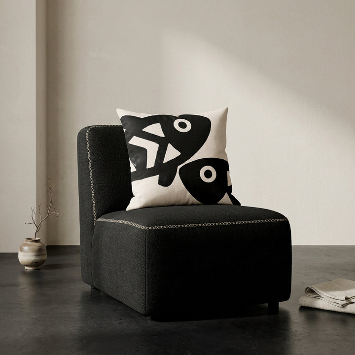 Photographie professionnelle qualité studio, d'un coussin carré mzelle dsign motifs poissons disposé sur une chaise japonaise sans accoudoirs, dossier en tissu noir surpiqué de fil écru, structure épaisse, disposé dans décor écru neutre, sol en béton ciré noir.