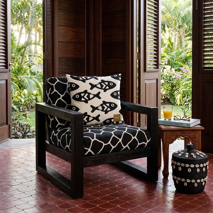 Photographie intérieure de style éditorial d’une villa créol,efauteuil épuré structure épaisse en bois noir et assise en lin noir, brodé de motifs blancs représentant des taches de girafes, orné d'un grand coussin carré mzelle design motifs poisson, placé contre un mur de panneaux de bois sombre. Au sol du carrelage rouge grenat en forme d'écaille de poisson, un panier noir décoré de caulis.