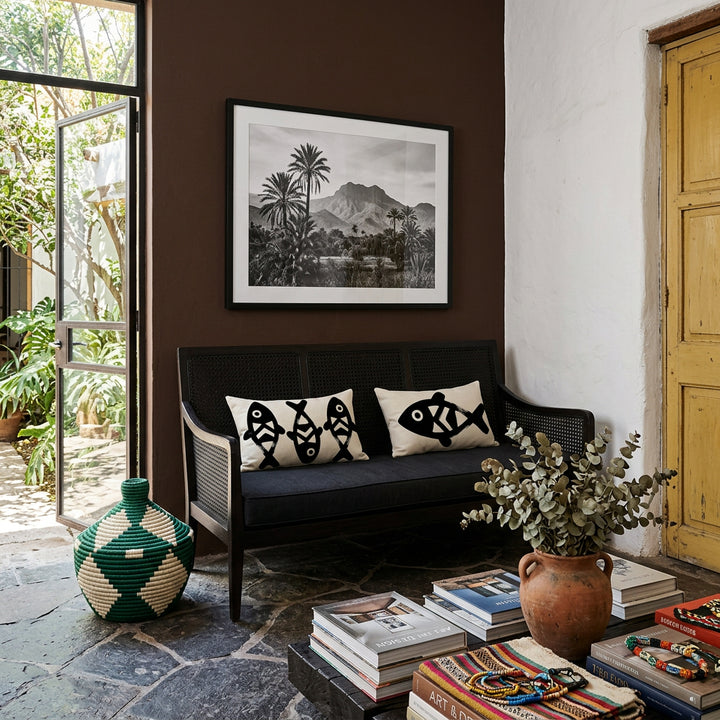 Photographie d’intérieur décoration d'hacienda modernisée. Canapé créole foncé au dossier en wengé et cannage noir, orné de deux coussins Mzelle design motifs poissons. Adossé à un mur peint en marron foncé uni sur lequel est accrochée une grande photographie en noir et blanc représentant un paysage exotique.