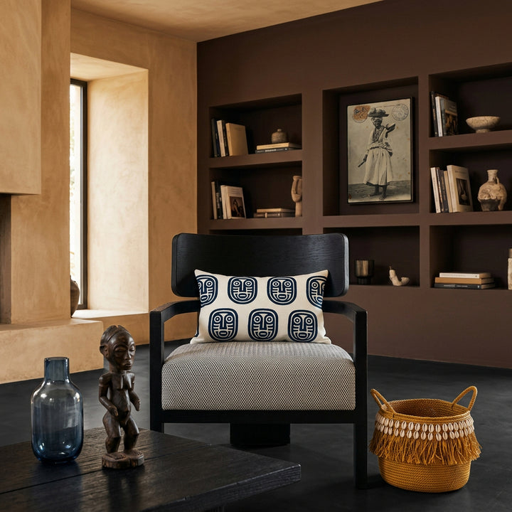 Photo professionnelle lifestyle, décoration Salon ethno-créole haut de gamme, chaise au design audacieux vue de face,  ornée d'un coussin rectangulaire mzelle design. Table basse en bois brûlé noir garni d'un vase en verre fumé bleu et d'une statuette style vaudou artistique.