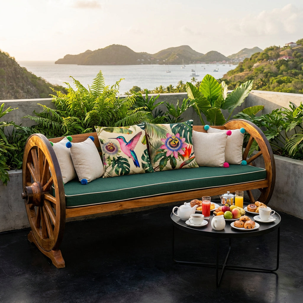 Photo professionnelle lifestyle roof top d'un hôtel aux Antilles, vue sur un paysage côtier nichée dans une baie,  un banc vu de face au design original avec 2 roues de charrettes aux extrémités, orné de 2 larges coussins mzelle design motif colibri et fleur de passiflore, une table basse ronde minimaliste assortie avec un plateau noir où est disposé un petit déjeuner élégant.