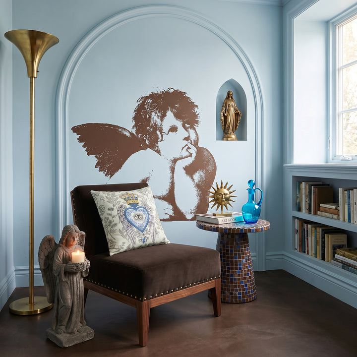 Photographie professionnelle décoration, fauteuil moderne orné d'un large coussin carré MZelle Design motif Ex Voto et herbier naturaliste, placé devant une fresque style graphique Pop Art représentant un angelot de Raphael, une Madone en laiton posée dans une niche, statue d'ange en pierre qui tient une bougie parfumée. Des Objets décoratifs sont disposés sur une table en mosaïque brun et bleu, sol en ciment brun foncé, grande lampe en laiton 1950 dans un coin. Ambiance invitante et intimiste.