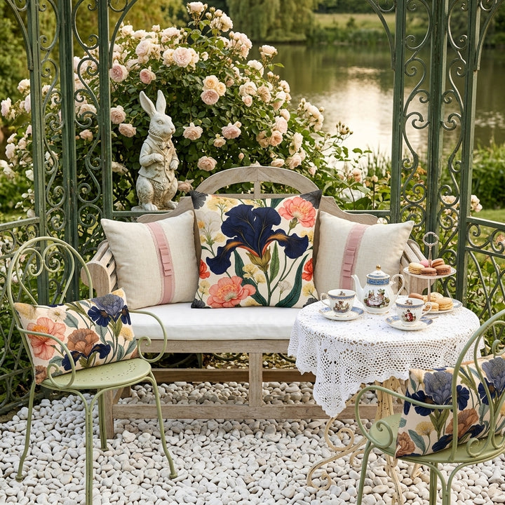 photographie professionnelle, décor extérieur Regencycore, Kiosque en fer forgé dans un jardin anglais , élégante banquette de jardin en bois clair et assise de coton blanc, orné d'un large coussin carré motif iris mzelle design et 2 autres en lin blanc , sur les côtés 2 chaises en fer forgé vert tilleul avec chacune un petit coussin rectangle parterre de galet blanc, table ronde avec un service à thé vintage. 