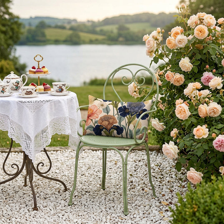 photographie professionnelle, décor extérieur Regencycore, une élégante chaise en fer forgé vert tilleul avec un petit coussin rectangle, parterre de galet blanc, table ronde avec un service à thé vintage en porcelaine fines motifs d’Alice au pays des merveilles et plateau de pâtisseries fines disposés sur une nappe en coton blanc ajourée, un buisson de roses anglaises, vue sur lac.