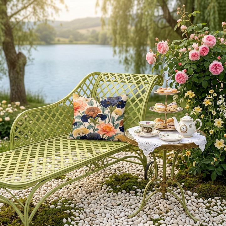 photographie professionnelle, décor extérieur Bridgerton, vue en plongée sur une élégante chaise en fer forgé vert tilleul avec un  coussin fleuri mzelle design, parterre de galet blanc, table ronde avec un service à thé vintage en porcelaine fines motifs d’Alice au pays des merveilles et plateau de pâtisseries fines disposés sur une nappe en coton blanc ajourée, un buisson de roses anglaises, vue sur lac.