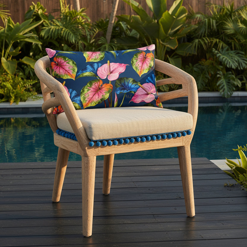 Photographie professionnelle d'un coussin mzelle design motifs anthuriums acidulés, disposé sur une chaise au design original et contemporain en bois flotté, assise écrue avec pompons bleu , placé devant une piscine foncé moderne , parterre de plantes exotiques et sol en bois noir sol. 