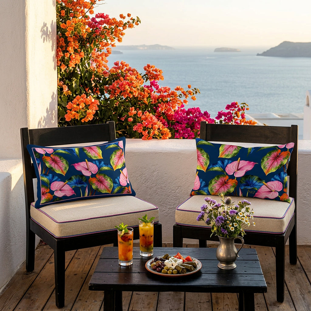 Photo professionnelle de décoration  hotel des cyclades, bougainvilliers orangé-fushia, scène baignée de soleil ,  2 fauteuils en bois foncé , assises écrues finitions des fines passementerie bleu et rose, chacun est orné d'un coussin rectangle mzelle design motifs anthuriums, bois au sol, table basse en bois noir avec 2 verres de jus de fruits, tapas, et un petit bouquet de fleurs sauvages.