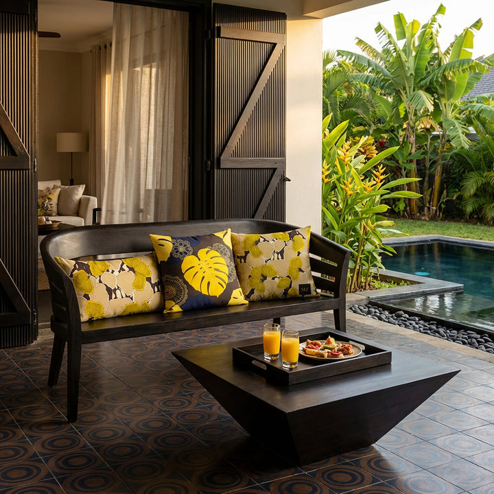 Photo lifestyle professionnelle décoration de villa créole avec une sublime banquette en ébène au design moderne, assise structure unie, ornée de trois coussins mzelle design, motifs floraux jaune et brun, table basse avec plateau contenant 2 verres de jus de fruit et une assiette de tapas. Portes en bois, aperçu d'un salon par de fins rideaux en lin, au sol un carrelage géométrique marron foncé et bleu marine. A côté d'un jardin tropical agrémenté d'une piscine moderne.