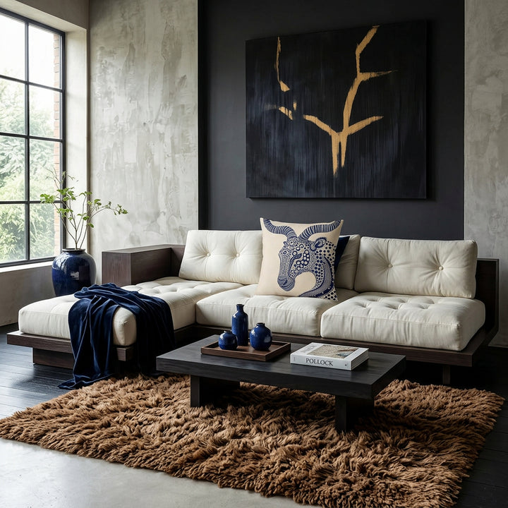 Photo professionnelle lifestyle esprit mix and match Belgian Minimalism, Japandi et Wabi-Sabi contemporain.
 grand canapé plateforme  en bois foncé et assises en twill ivoire capitonnés, orné de coussins mzelle design motif animalier stylisés. Murs minéraux texturés en enduit à la chaux et béton ciré gris perle, sublimés par une œuvre abstraite monumentale noire et or inspirée des galeries d'art contemporaines. Table basse monolithique en bois noirci, céramiques artisanales minimalistes bleu outremer. 