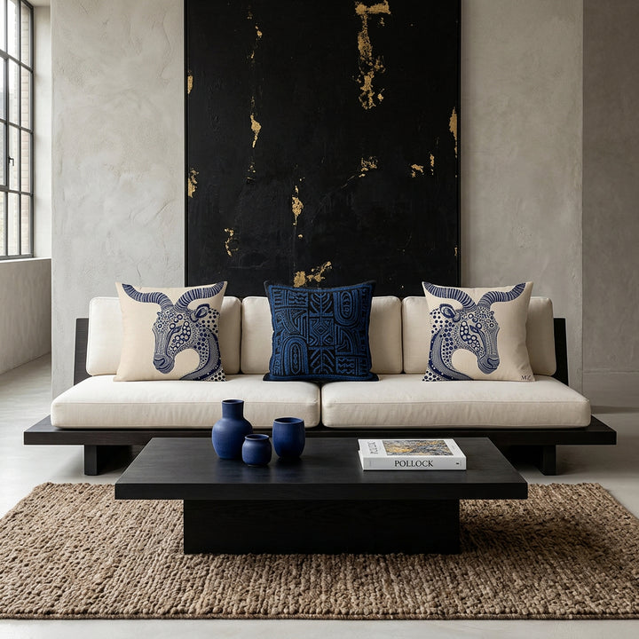 Photo professionnelle lifestyle esprit mix and match Belgian Minimalism, Japandi et Wabi-Sabi contemporain.
 grand canapé plateforme  en bois foncé et assises en twill ivoire capitonnés, orné de coussins mzelle design motif animalier stylisés. Murs minéraux texturés en enduit à la chaux et béton ciré gris perle, sublimés par une œuvre abstraite monumentale noire et or inspirée des galeries d'art contemporaines. Table basse monolithique en bois noirci, céramiques artisanales minimalistes bleu outremer. 