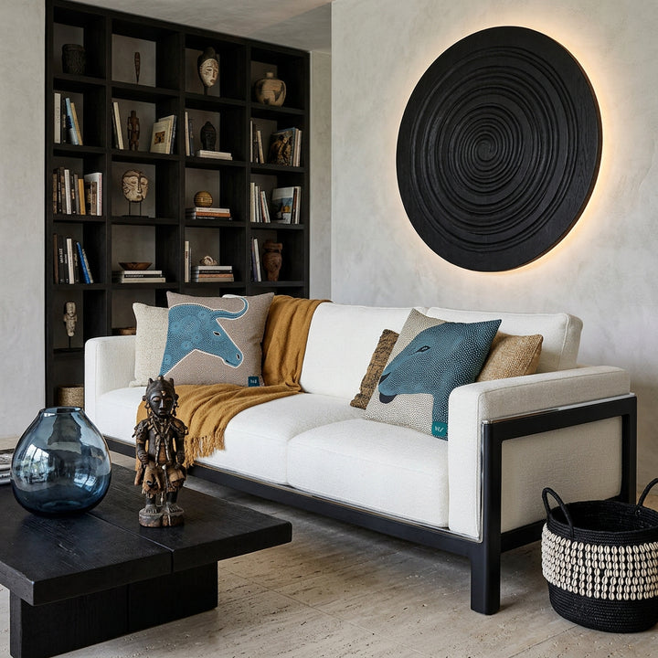 Photo professionnelle lifestyle, décoration Salon ethno-brutaliste haut de gamme, imposant canapé en lin  structure épaisse en polypropylène noir, liseré chrome , orné de 2 larges coussins graphiques mzelle design motif graphique animaux , table basse en bois brûlé noir garni d'un vase en verre fumé bleu et d'une statuette style vaudou artistique, architecture minérale et mur  enduit à la chaux texturé avec une bibliothèque occupant tout le mur et une grande applique lumineuse.