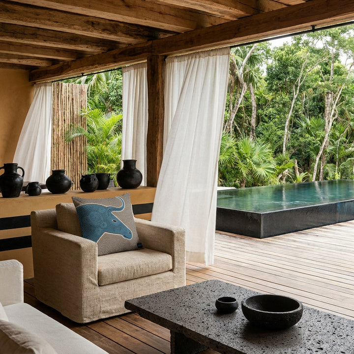Photo professionnelle lifestyle, décoration terrasse d’eco-lodge tropical haut de gamme, fauteuil en tissu naturel écru texturé orné de d'un coussin mzelle design motif graphique , table basse en pierre volcanique, sol en bois brut, poteries artisanales sur un muret peint de rayures noires, rideaux en lin flottant dans une brise chaude, architecture ouverte sur une jungle luxuriante et piscine sombre moderne.