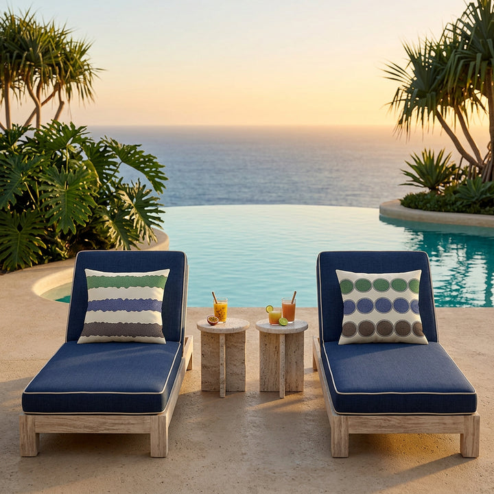 Photo professionnelle lifestyle décoration villa beach side contemporaine inspirée de la Polynésie moderne, vue de face sur deux chaises longues bleu indigo, structure épaisse en bois flotté blanchi, orné de coussins mzelle design motifs géométriques, sol travertin clair, terrasse ouverte sur l’océan, béton minéral sableux, végétation tropicale graphique maîtrisée, lumière dorée de coucher de soleil. Ambiance séjour en vacances. 