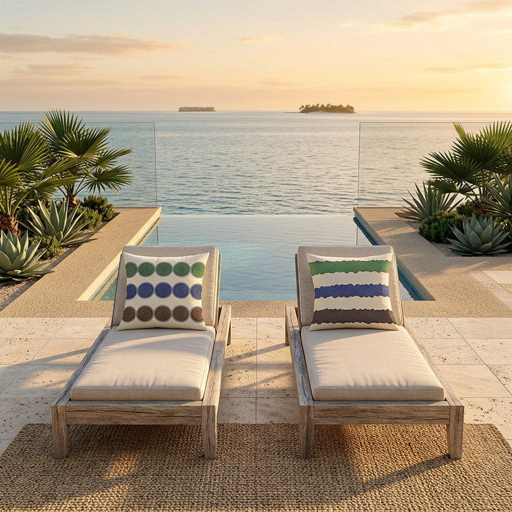 Photo professionnelle lifestyle décoration villa beach side contemporaine inspirée de la Polynésie moderne, vue de face sur deux chaises longues, structure épaisse en bois flotté blanchi, orné de coussins mzelle design motifs géométriques, sol travertin clair, terrasse ouverte sur l’océan, béton minéral sableux, végétation tropicale graphique maîtrisée, lumière dorée de coucher de soleil. Ambiance séjour en vacances. 