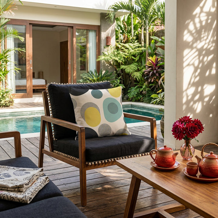 Photo professionnelle de décoration cour intérieure d'une maison d'hôte à Bali,  fauteuil moderne en bois de manguier assises lin noires finitions petits pompons écrus, orné d'un large coussin mzelle design motifs vintage géométriques, dalles de caillebotis en bois au sol, table basse assortie avec un service à thé en grès rouge dégradé et un petit bouquet de dahlias rouges dans un vase en laiton, en arrière plan jardin tropical et piscine . 