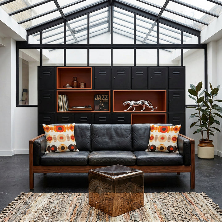 photo de décoration, Grand loft industriel, Mid-Century Modern hybride et éclectique, avec verrière noire style atelier baigné de lumière naturelle, vue de face sur un canapé vintage en cuir noir orné de 2 coussins mzelle design motifs géométriques retro. En arrière plan: Bibliothèque modulaire asymétrique noir mat avec niches orange brûlé en résine, style anciens casiers industriels, une Platine et des vinyles, livre de jazz. 