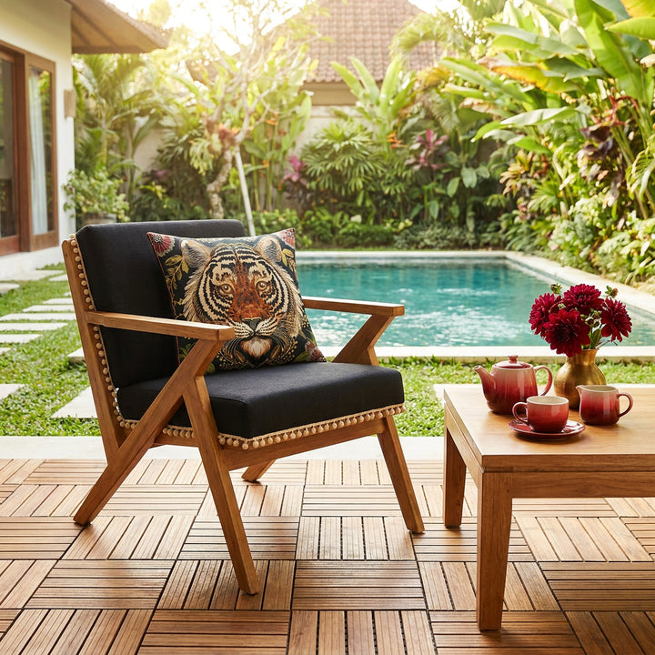 Photo professionnelle, scène baignée de soleil,  fauteuil moderne en bois de manguier structure en V et assises lin noires finitions petits pompons écrus, orné d'un large coussin mzelle design motif tigre indien, dalles de caillebotis en bois au sol, table basse assortie avec un service à thé en grès rouge dégradé et un petit bouquet de dahlias, arrière plan jardin tropical et piscine . 