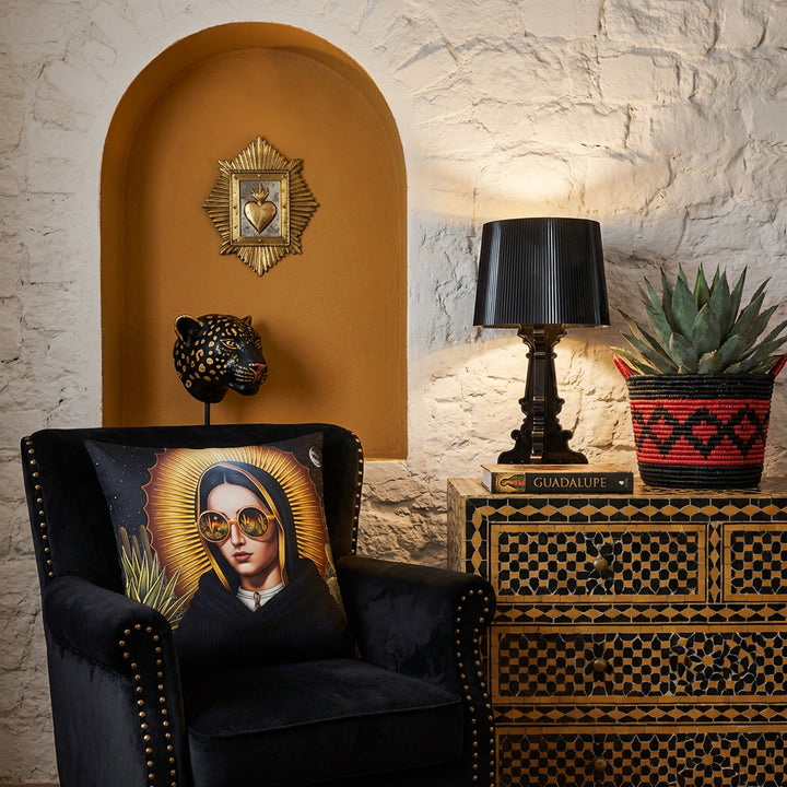 Photographie professionnel décoration  loft mexicain, chaise en velours noir finitions rivets laiton design moderne, orné d'un large coussin mzelle design motif vierge marie, placé devant une niche en arche ocre dans le mur en pierres blanchies, décoré d'un EX VOTO en laiton et d'une tête de jaguar en peinture noir. 