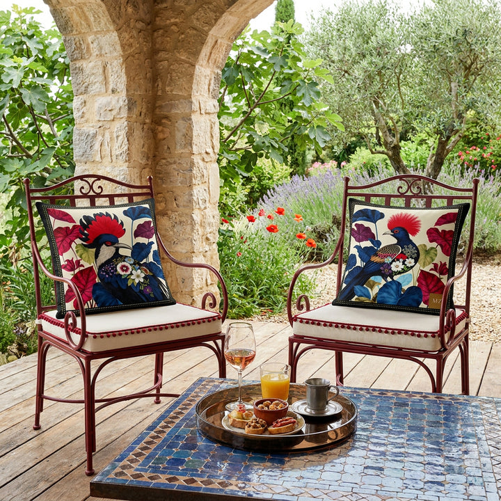 Photo professionnelle de décoration d'intérieur, terrasse d'une villa de la côte d'azur baignée de soleil , 2 chaise en fer forgé rouge grenat, assises écru finitions fins pompons rouge grenat, orné de chaque côté d'un coussin mzelle design motif oiseaux et caladium , dalles de bois rustique au sol, table basse en mosaiques bleu et brun, plateau rond en plexiglass brun fumé.
