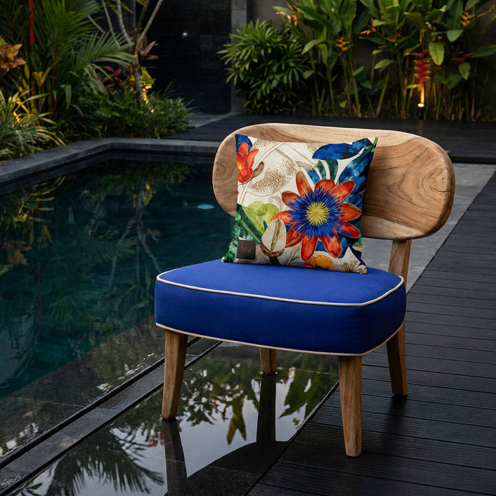 Photographie professionnelle d'un coussin carré mzelle design motif fleurs tropicales, disposé sur une chaise au design original et contemporain en bois flotté, assise bleu outremer finitions piping écru , placé devant une piscine foncé moderne , parterre de plantes exotiques et sol en bois noir sol. 