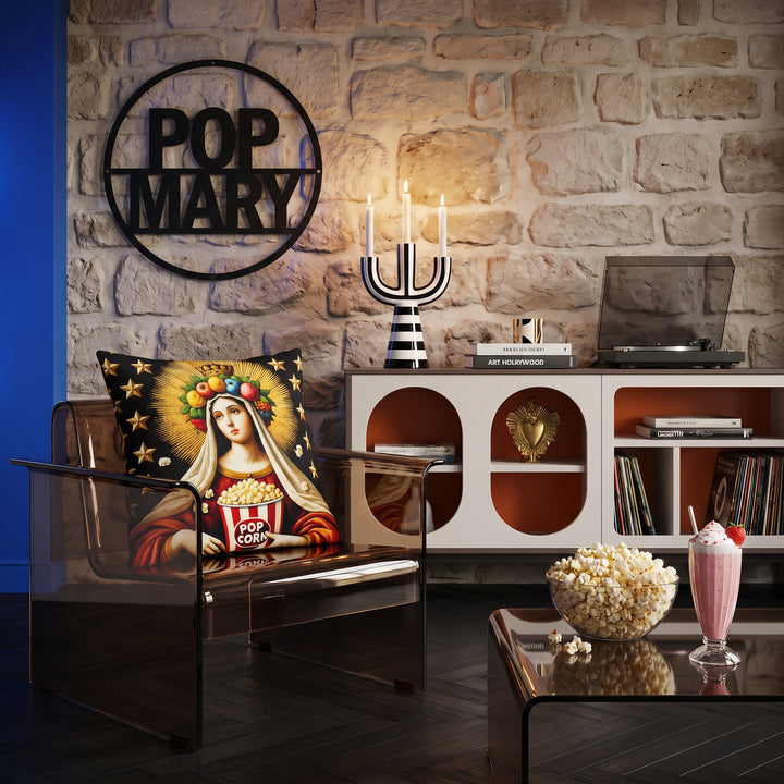 Photographie d'un salon qui fusionne style pop art, vue de face d'un fauteuil en plexiglas brun fumé au design audacieux, orné d'un large coussin mzelle design motif vierge marie humour, mur en pierres où est accroché un lettrage "POP MARY" métal noir, table basse assortie avec un bol de pop corn et un milk shake fraise.