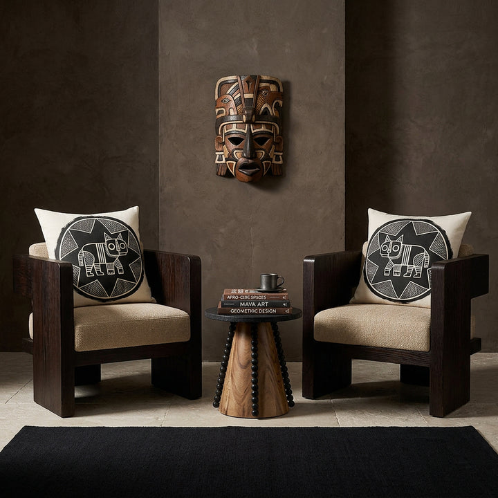 Photo professionnelle de décoration d'intérieur, salon afro chic brutaliste, deux fauteuils sculpturaux bas en bois de wengé massif aux lignes géométriques monolithiques, assises tissu texturé sable, chacun est orné d'un large coussin mzelle design motif félin noir et écru . Murs en béton brut teinté argile sombre décoré d'un masque maya en bois peint . Table basse artistique avec un socle en bois et perles noires.