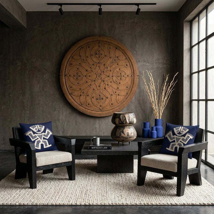 Photo professionnelle de décoration d'intérieur, salon brutaliste précolombien, vue de face. deux fauteuils sculpturaux bas en bois de noir vernis aux lignes géométriques monolithiques, assises tissu texturé écru, chacun est orné d'un large coussin mzelle design bleu et écru . Murs en béton brut teinté argile sombre décoré d'un bouclier en bois peint . Table basse monolithique en pierre volcanique.
