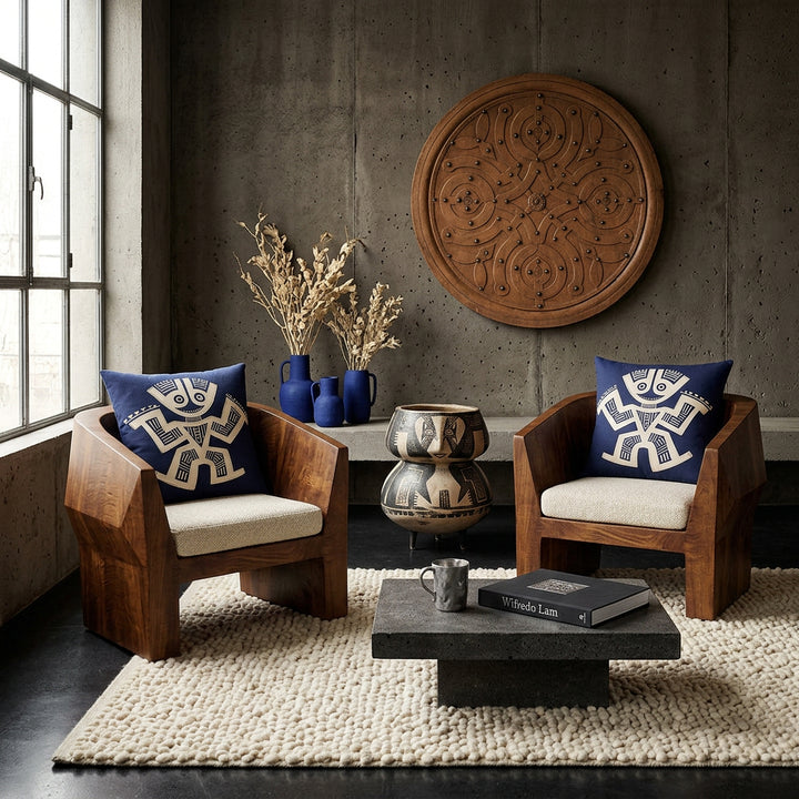 Photo professionnelle de décoration d'intérieur, salon brutaliste précolombien, vue de face. deux fauteuils sculpturaux bas en bois de manguier vernis aux lignes géométriques monolithiques, assises tissu texturé écru, chacun est orné d'un large coussin mzelle design bleu et écru . Murs en béton brut teinté argile sombre décoré d'un bouclier en bois peint . Table basse monolithique en pierre volcanique.